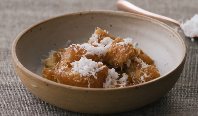 A ceramic bowl containing golden-brown fried pastry pieces topped with shredded white coconut on a gray woven fabric.